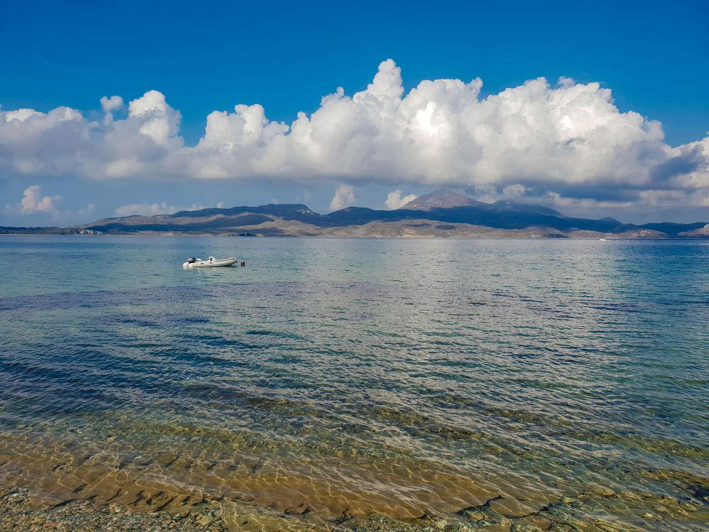 32168358 Serene view of a boat floating on the Aegean Sea with Milos Island in the background under a clear sky.